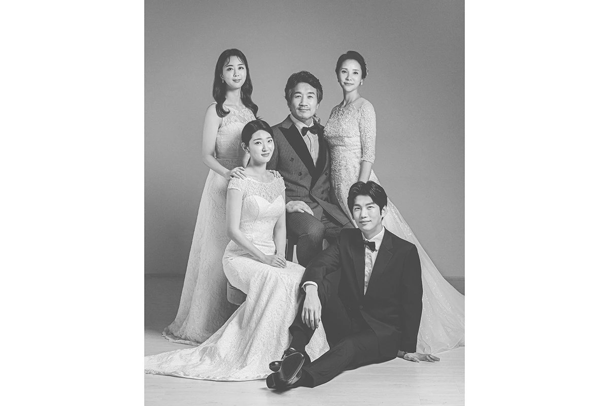 밝은 흰색 스튜디오에서 턱시도와 드레스를 입은 성인 5명이 흰 배경 앞 의자에 앉고 선 사진이며, 의자와 부케가 장면의 구성을 보완합니다.