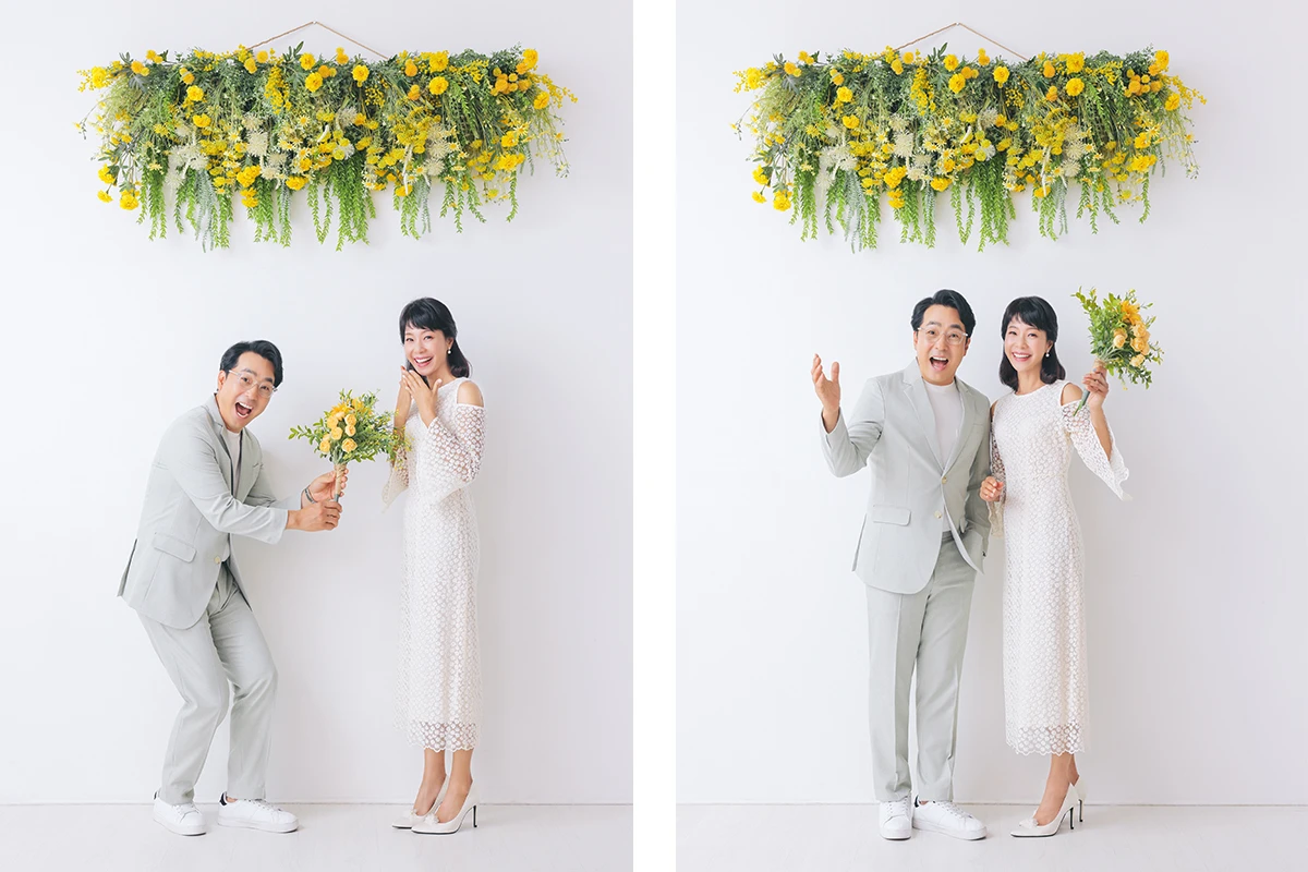 밝은 창가와 분홍 꽃 장식에서 턱시도와 드레스를 입은 성인 2명이 꽃 장식 사이에 앉아 있는 사진이며, 꽃 장식과 넓은 드레스 자락가 장면의 구성을 보완합니다.