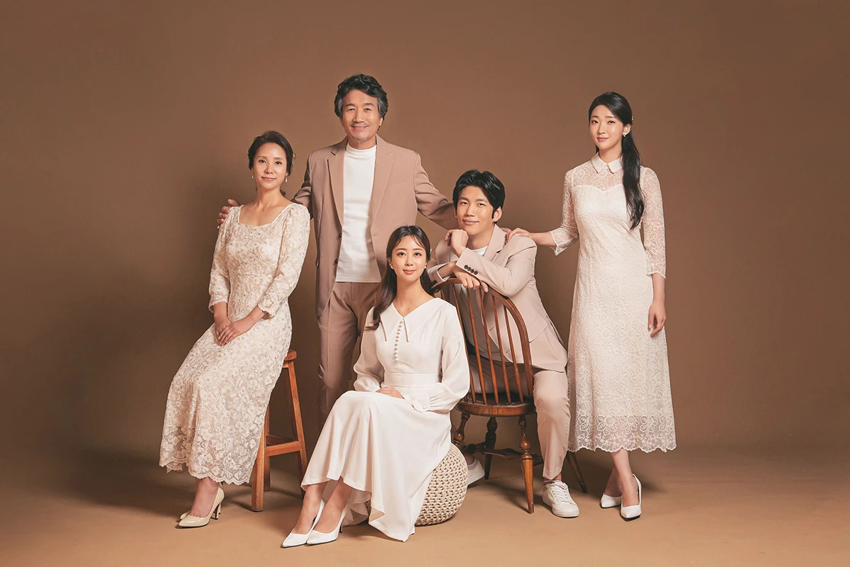 밝은 흰색 스튜디오에서 베이지 정장과 흰 원피스의 성인 2명이 흰 배경과 노란 꽃 장식 아래 서 있는 분할 사진이며, 노란 꽃 장식과 부케가 장면의 구성을 보완합니다.