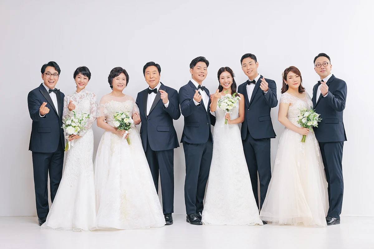 회색 배경과 검정 소파가 있는 스튜디오에서 턱시도와 화이트 드레스를 입은 성인 2명이 검정 소파 앞에 앉은 상반신 사진이며, 검정 소파와 포멀 의상가 장면의 구성을 보완합니다.