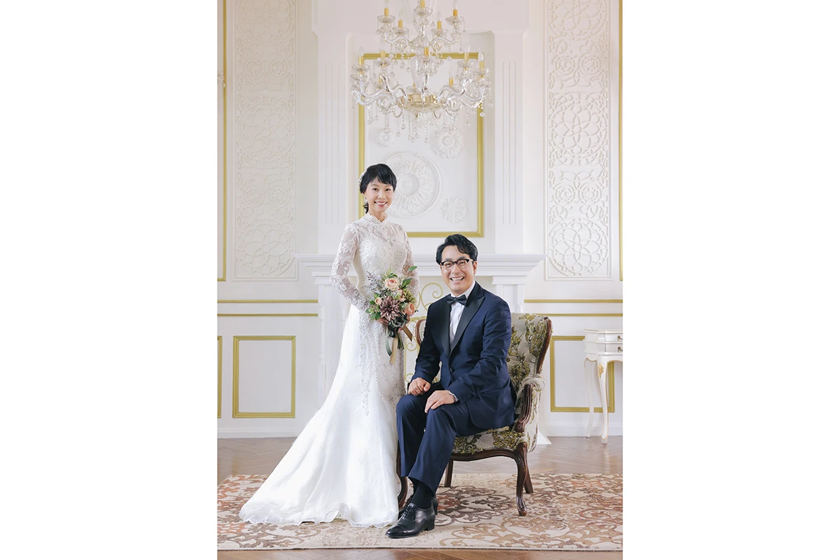 샹들리에와 몰딩이 있는 클래식 실내에서 턱시도와 화이트 드레스를 입은 성인 2명이 클래식 실내 세트에 배치된 사진이며, 의자, 부케, 러그가 장면의 구성을 보완합니다.