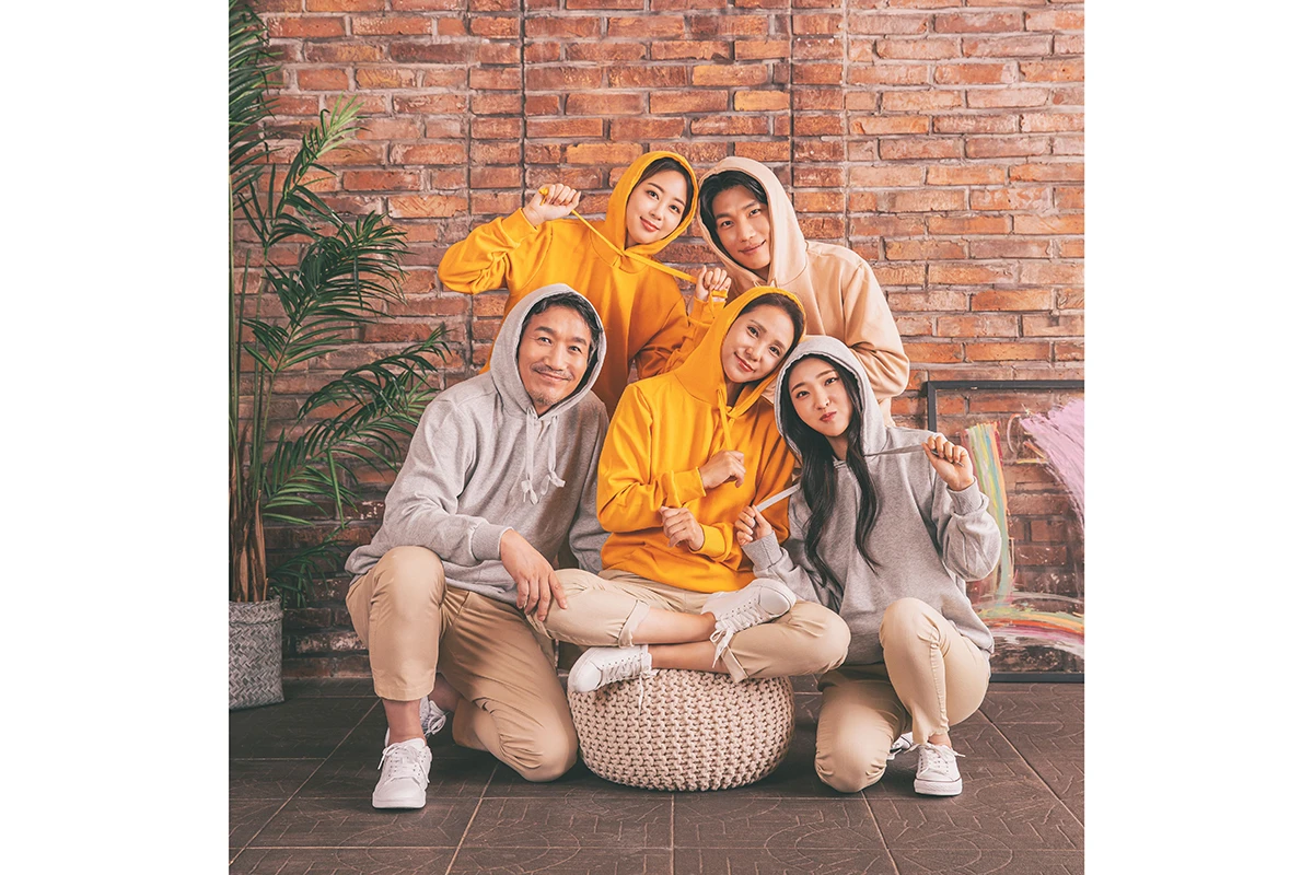 밝은 흰색 스튜디오에서 후드 티셔츠와 캐주얼 팬츠를 입은 성인 5명이 밝은 흰 배경 앞에 앉은 사진이며, 노란 후드와 회색 후드 의상가 장면의 구성을 보완합니다.
