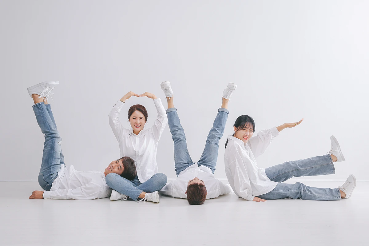 밝은 흰색 스튜디오에서 교복풍 의상과 검정 정장을 입은 성인 4명이 흰 배경 앞에서 역할극처럼 배치된 사진이며, 가방, 모자, 교복풍 의상가 장면의 구성을 보완합니다.