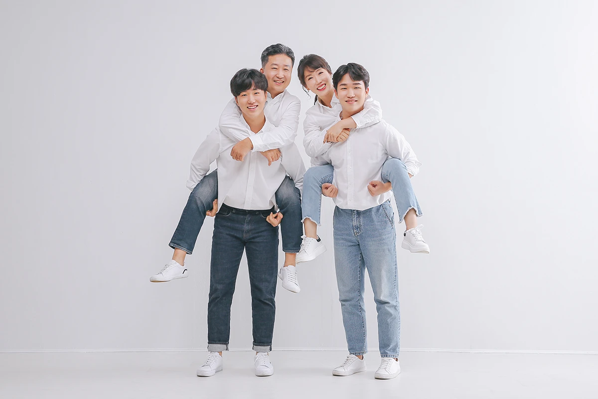벽돌 벽과 회색 소파가 있는 실내에서 흰 셔츠와 청바지를 입은 성인 3명과 흰 반려견이 벽돌 배경 앞 소파에 앉은 사진이며, 반려견, 소파, 식물가 장면의 구성을 보완합니다.